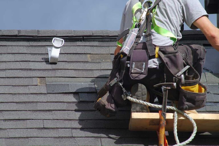 Roofing workers inspect a residential roof deck beside stacked materials and protective ground coverings.